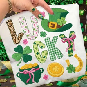 May include: White fabric with the word "LUCKY" in various patterns and colors, including leopard print, pink floral, green gingham, and green shamrocks. The design also features a green leprechaun hat, shamrocks, a pot of gold, a gold coin, and a horseshoe.