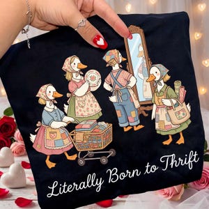 May include: Black t-shirt featuring a whimsical illustration of anthropomorphic ducks dressed in vintage clothing, engaged in thrifting activities. The phrase "Literally Born to Thrift" is written in a cursive font at the bottom of the shirt.