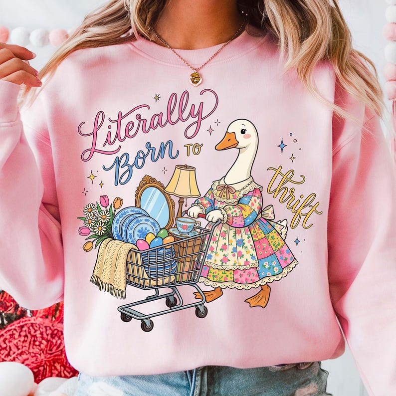 Może przedstawiać: R&oacute;żowa bluza z zabawną ilustracją gęsi pchającej w&oacute;zek sklepowy wypełniony przedmiotami vintage. Gęś ma na sobie patchworkową sukienkę, a napis brzmi "Literally Born to Thrift".