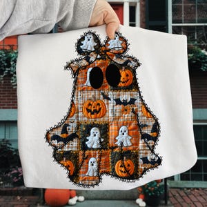 Può includere: Un cuscino decorativo bianco a tema Halloween a forma di fantasma. Il design del fantasma presenta un motivo patchwork con zucche, pipistrelli e figure di fantasmi in arancione, nero e bianco. I bordi sono delineati con cuciture nere.