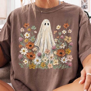 May include: A brown short-sleeve t-shirt featuring a central graphic of a white ghost surrounded by a variety of colorful flowers including orange poppies, white daisies, and pink clover blossoms, rendered in a textured, painterly art style.