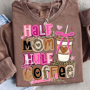 May include: A brown sweatshirt with the text "Half Mom Half Coffee" in pink, brown, and black. The design includes a coffee cup with a pink bow and a heart, along with star and leopard print accents. A silver necklace is visible.