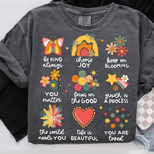 May include: A dark gray t-shirt with a colorful graphic design featuring flowers, butterflies, rainbows, and stars. The graphic includes the text "be kind always", "choose joy", "keep on blooming", "you matter", "focus on the good", "growth is a process", "the world needs you", "life is beautiful", and "you are loved".