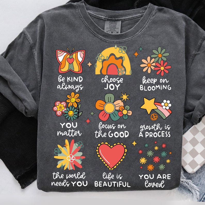 May include: A dark gray t-shirt with a colorful graphic design featuring flowers, butterflies, rainbows, and stars. The graphic includes the text "be kind always", "choose joy", "keep on blooming", "you matter", "focus on the good", "growth is a process", "the world needs you", "life is beautiful", and "you are loved".
