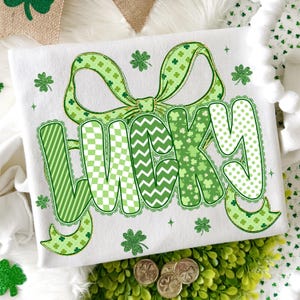 Puede incluir: Camiseta blanca con un gran lazo verde y la palabra "LUCKY" en varios patrones verdes. Las letras est&aacute;n decoradas con rayas, chevrones y lunares. Acentos de tr&eacute;boles y monedas de oro completan el tema del D&iacute;a de San Patricio.