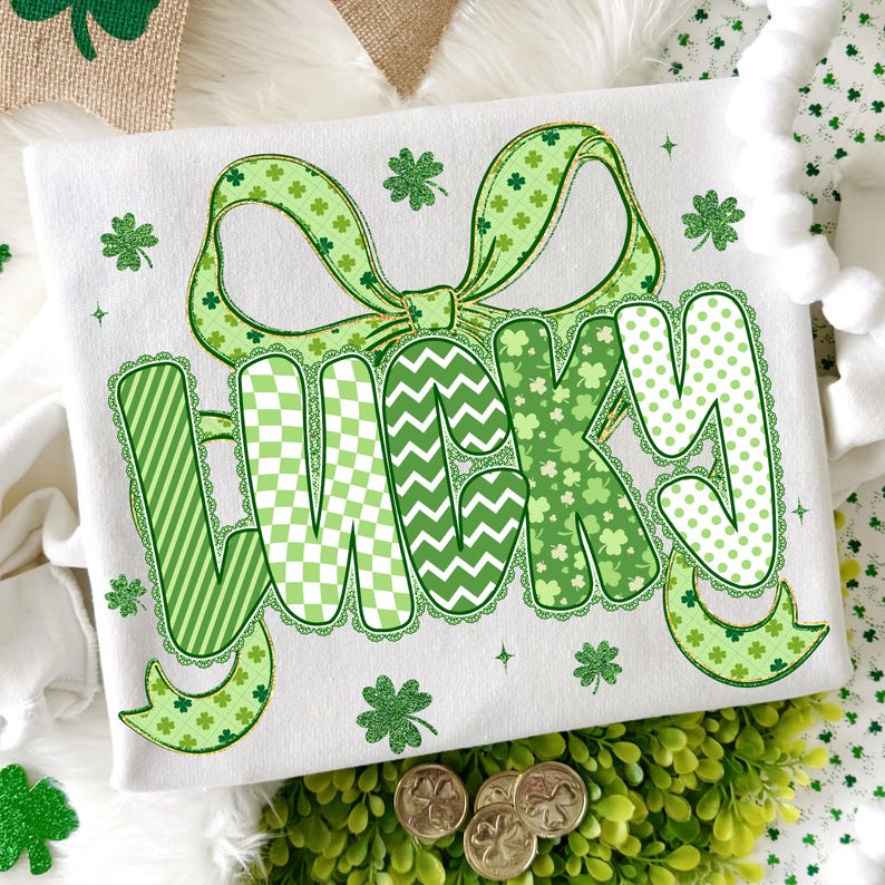 Puede incluir: Camiseta blanca con un gran lazo verde y la palabra "LUCKY" en varios patrones verdes. Las letras est&aacute;n decoradas con rayas, chevrones y lunares. Acentos de tr&eacute;boles y monedas de oro completan el tema del D&iacute;a de San Patricio.