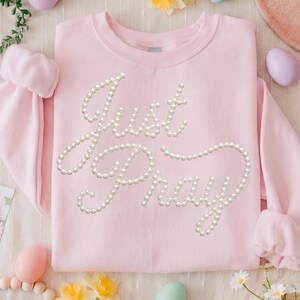 Op de afbeelding: Een lichtroze sweatshirt met de woorden "Just Pray" uitgespeld in imitatieparel kralen. De trui heeft een ronde hals en lange mouwen. De achtergrond bevat pastelkleurige paaseieren en bloemen.