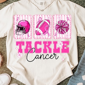 May include: Cream-colored t-shirt with a pink graphic supporting cancer awareness. The design includes a football helmet, football, and pom-pom, each with a pink ribbon. The words "TACKLE Cancer" are in pink.