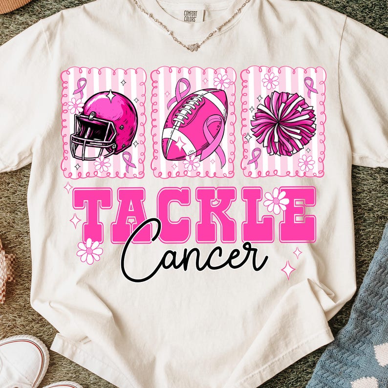 May include: Cream-colored t-shirt with a pink graphic supporting cancer awareness. The design includes a football helmet, football, and pom-pom, each with a pink ribbon. The words "TACKLE Cancer" are in pink.