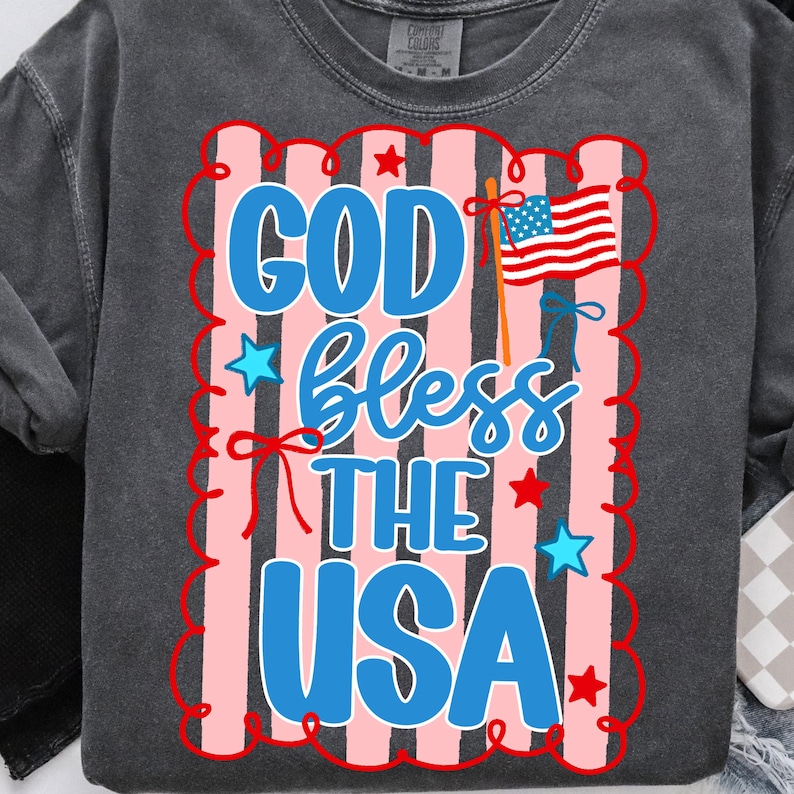 Puede incluir: Una sudadera gris con un dise&ntilde;o patri&oacute;tico. El dise&ntilde;o presenta el texto "GOD bless THE USA" en azul y blanco, con una peque&ntilde;a bandera estadounidense y estrellas rojas. El fondo tiene rayas rosas y blancas.