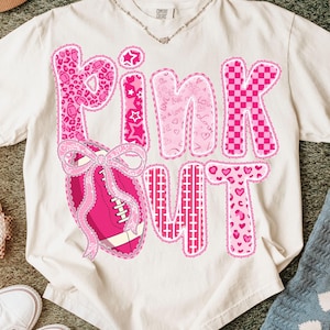 May include: Cream-colored t-shirt with the words "PINK OUT" in pink, featuring various patterns like leopard print, stars, hearts, and a football with a bow. The shirt has a crew neck and short sleeves.