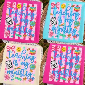 May include: Four t-shirts with the text "Teaching is my ministry" and school-themed graphics. The shirts are pink, light blue, cream, and pink, each with a different background colour. The design includes a globe, books, and a notebook.