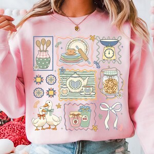 May include: Pink sweatshirt with a whimsical kitchen-themed design. The graphic includes illustrations of kitchen tools, dishes, a teapot, cookies, and ducks. The design is framed in decorative borders with floral accents.