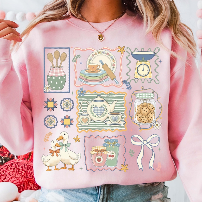 May include: Pink sweatshirt with a whimsical kitchen-themed design. The graphic includes illustrations of kitchen tools, dishes, a teapot, cookies, and ducks. The design is framed in decorative borders with floral accents.