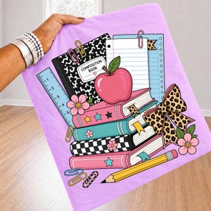 May include: A light purple fabric featuring a school-themed graphic print. The design includes a black and white composition book, a lined notepad, a red apple, a stack of colorful books, a ruler, a yellow pencil, paper clips, and a leopard print bow.