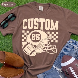 May include: A brown short-sleeve t-shirt featuring a cream-colored graphic of a football helmet with the number 25, a football, and a checkered pattern. The word CUSTOM is printed in large capital letters above the helmet graphic.