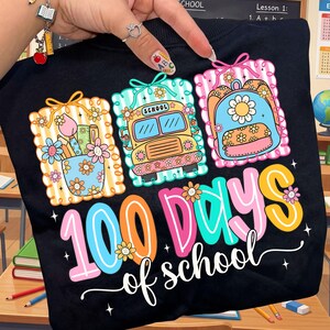 K&ouml;nnte beinhalten: Schwarzes T-Shirt mit einem farbenfrohen Design zur Feier von "100 Schultagen". Das Design zeigt einen Schulbus, einen Rucksack und eine Tasse mit Bleistiften und Blumen. Der Text "100 Schultage" ist in einer verspielten Schriftart mit floralen Akzenten.