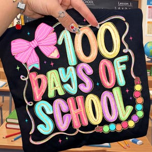 Pode incluir: Camiseta preta com um gráfico colorido celebrando "100 dias de escola". O design apresenta um grande laço rosa, um lápis e o texto em várias cores com detalhes de glitter dourado. A camiseta é segurada por uma mão.