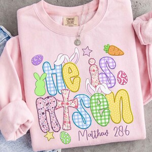 May include: A light pink Comfort Colors sweatshirt with the words "He is Risen" in colorful, patterned letters. Easter-themed graphics include a cross, eggs, a carrot, and a bunny. The text "Matthew 28:6" is printed below.