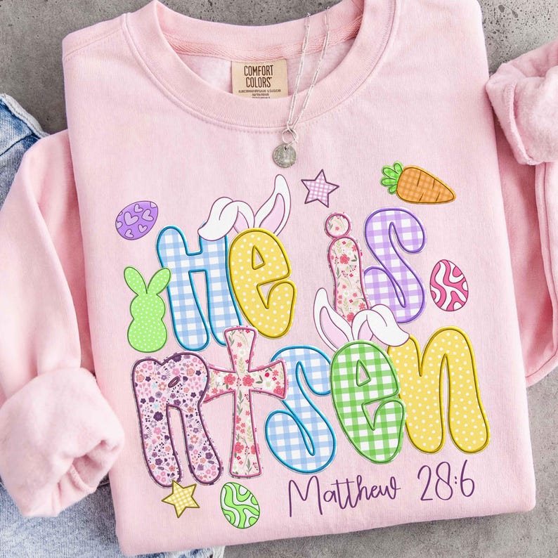 May include: A light pink Comfort Colors sweatshirt with the words "He is Risen" in colorful, patterned letters. Easter-themed graphics include a cross, eggs, a carrot, and a bunny. The text "Matthew 28:6" is printed below.