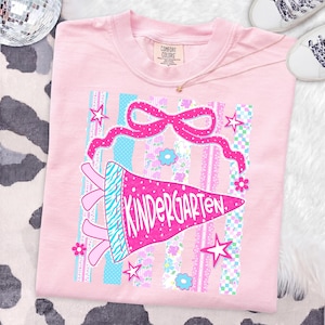 May include: A light pink t-shirt with a colorful graphic design. The design features a pink pennant that says "KINDERGARTEN" in white, a pink bow, stars, and floral accents. The shirt is laid flat on a cow-print surface.