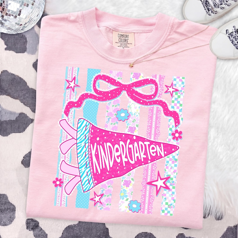 May include: A light pink t-shirt with a colorful graphic design. The design features a pink pennant that says "KINDERGARTEN" in white, a pink bow, stars, and floral accents. The shirt is laid flat on a cow-print surface.