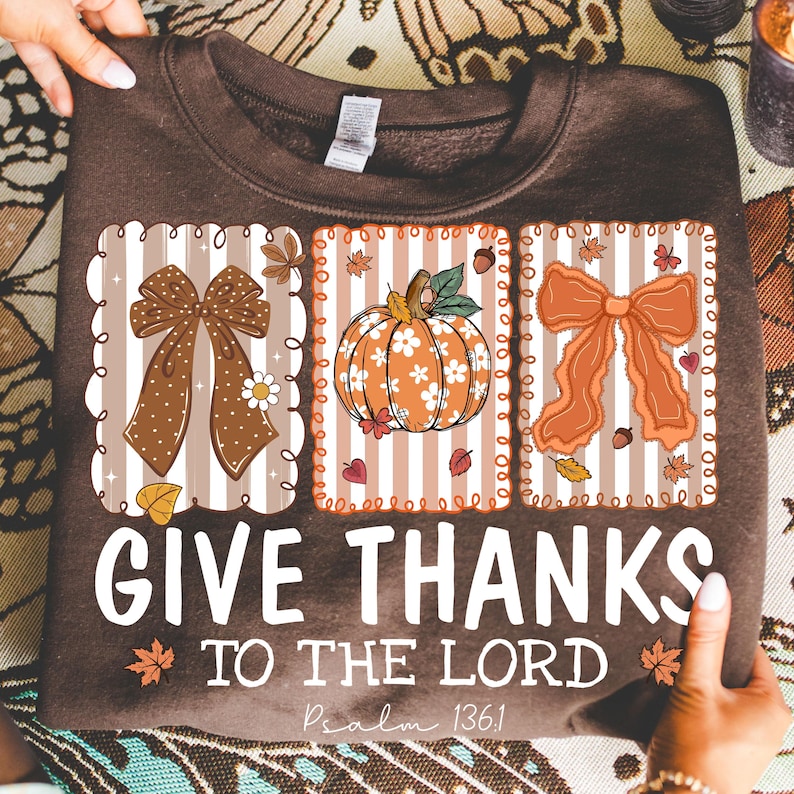 May include: A brown sweatshirt with a fall-themed design. The design includes three rectangular panels with a bow, a pumpkin, and another bow. The text "GIVE THANKS TO THE LORD" is printed below the panels, with "Psalm 136:1" underneath.