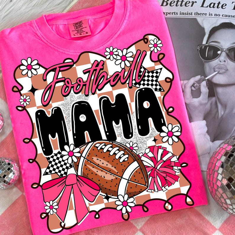 May include: A bright pink t-shirt with the words "Football MAMA" in a playful font. The design includes a football, daisies, a bow, and a pom-pom. The shirt has a crew neck and short sleeves.