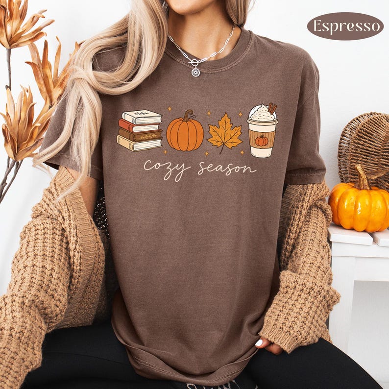 May include: A brown t-shirt featuring a graphic print of a stack of books, an orange pumpkin, a maple leaf, and a coffee cup with whipped cream. The text "cozy season" is printed below the graphics in a white script font. The shirt has a crew neckline.