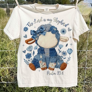 Puede incluir: Camiseta color crema con un diseño de cordero de retazos con detalles de mezclilla azul y flores. El texto "The Lord is my Shepherd" y "Psalm 23:1" están impresos en la camiseta. La camiseta está colgada en un tendedero.