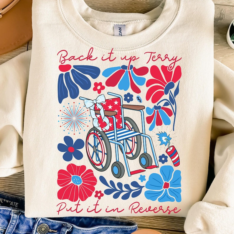 May include: Cream-colored sweatshirt with a patriotic design. The graphic features a wheelchair decorated with stars and stripes, surrounded by red and blue flowers and fireworks. Text reads "Back it up Terry, Put it in Reverse."