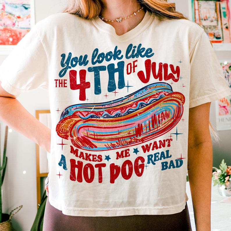 May include: Cream-colored t-shirt with a graphic of a hot dog and text that reads "You look like the 4th of July makes me want a hot dog real bad." The design features red, blue, and white colors, perfect for Independence Day celebrations.