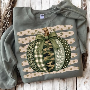 Op de afbeelding: Een saliegroene sweater met ronde hals en een grote afbeelding van een pompoen met een strik. De pompoen heeft segmenten met luipaardprint, groen gingham, camouflage, witte bladpatronen en stippen, tegen een achtergrond van beige strepen.