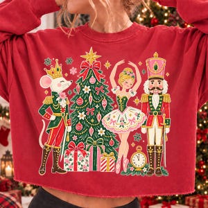 May include: A red long-sleeve sweatshirt featuring a graphic print of a mouse in a green uniform, a decorated Christmas tree, a ballerina in a white dress, and a nutcracker soldier, all surrounded by small snowflakes and a gold clock.