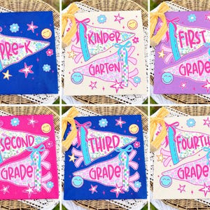 May include: A collection of six square fabric signs featuring pennant designs with school grade levels from Pre-K to Fourth Grade. Each sign displays colourful floral and checkered patterns, smiley faces, and stars, accented with a yellow ribbon.
