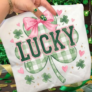 Könnte beinhalten: Weißes Kissen mit grünem und rosa Design. Das Kissen zeigt ein großes grün-weiß kariertes Kleeblatt mit dem Wort "LUCKY" in grünen Buchstaben, die rosa umrandet sind. Eine rosa Schleife und rosa Herzen verzieren das Kissen.