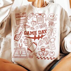 May include: Cream-colored sweatshirt with a red graphic design featuring football-related icons and the words "GAME DAY." The design includes a football, helmet, trophy, and other sports-themed elements.