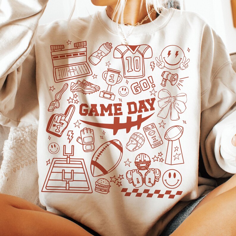 May include: Cream-colored sweatshirt with a red graphic design featuring football-related icons and the words "GAME DAY." The design includes a football, helmet, trophy, and other sports-themed elements.