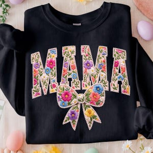 May include: Black sweatshirt featuring the word "MAMA" in large, floral letters. The letters are filled with colourful flowers and outlined in white. A large bow, also filled with flowers, is positioned below the word.