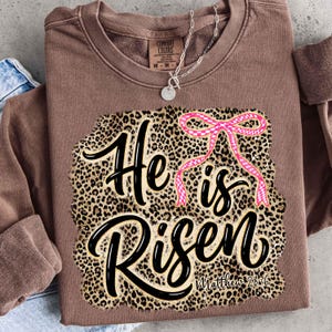 May include: A brown long-sleeved sweatshirt featuring the phrase "He is Risen" in black script, set against a leopard print design. A pink bow is above the word "is". The text "Matthew 28:6" is at the bottom.