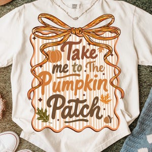 May include: Cream-colored t-shirt with a fall-themed graphic. The design features the text "Take me to The Pumpkin Patch" in brown and orange, with a pumpkin, leaves, and a bow. The shirt is made of a soft material.