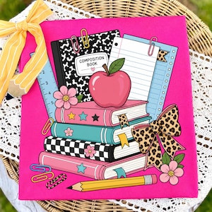 May include: A bright pink fabric featuring a graphic illustration of a stack of four books, a composition book, a red apple, a yellow pencil, a ruler, and colorful paper clips. A large leopard print bow and pink flowers decorate the school-themed design.