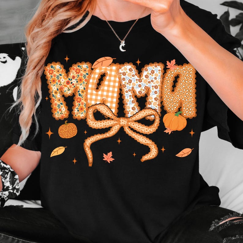 May include: Black t-shirt with the word "MAMA" in orange and floral patterns. A large bow and pumpkins are below the word. Autumn leaves and stars are scattered around the design.