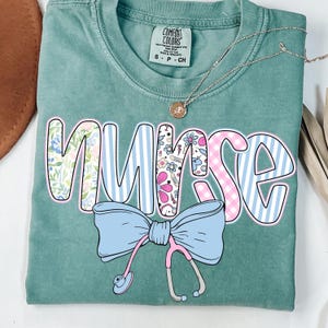 May include: A teal blue t-shirt with the word "NURSE" in pink, blue, and white patterned letters. A blue bow with a pink stethoscope is below the word. The shirt has a tag that says "COMFORT COLORS".