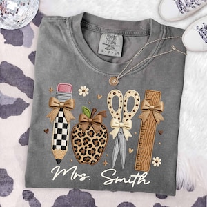 May include: A gray t-shirt featuring an embroidered design of a checkered pencil, a leopard print apple, a pair of scissors, and a wooden ruler, each accented with tan bows. The text Mrs. Smith is embroidered in white script below the school supplies.