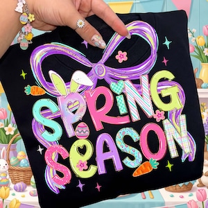 May include: Black sweatshirt with a colorful "Spring Season" graphic. The design features a large purple bow, bunny ears, carrots, and Easter eggs. The text is in various colors, including pink, green, and blue, with decorative patterns.