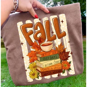 Op de afbeelding: Een bruine tote bag met een grafische print met het woord Fall in grote bruine letters. Daaronder staat een kopje thee op een stapel boeken met de titels Crisp Air, Cozy Days, Autumn Stories, Pumpkin Spice en Sweater Weather, met herfstbladeren.