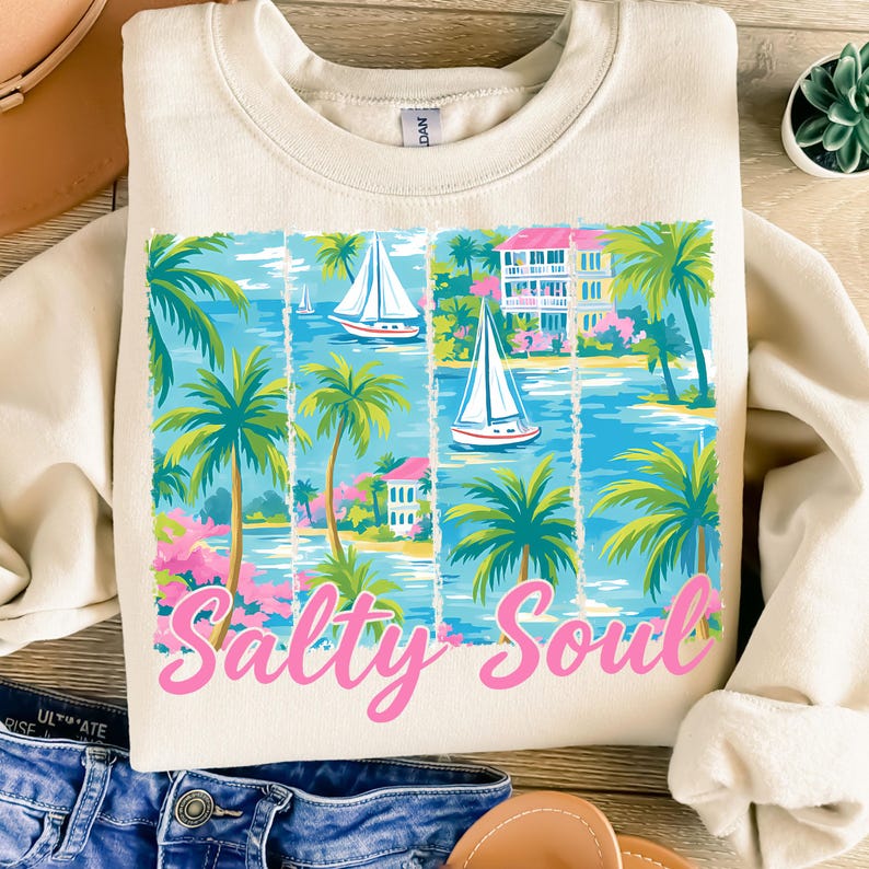 K&ouml;nnte beinhalten: Ein beiger Pullover mit einem grafischen Druck einer tropischen Szene mit Palmen, Segelbooten und H&auml;usern. Der Text "Salty Soul" ist in Pink unter dem Grafikdruck gedruckt.