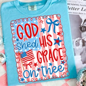 May include: A light blue t-shirt with a patriotic design. The graphic features the text "GOD SHED HIS GRACE ON THEE" in red and blue, along with an American flag and stars. The design is framed with a red and white border.