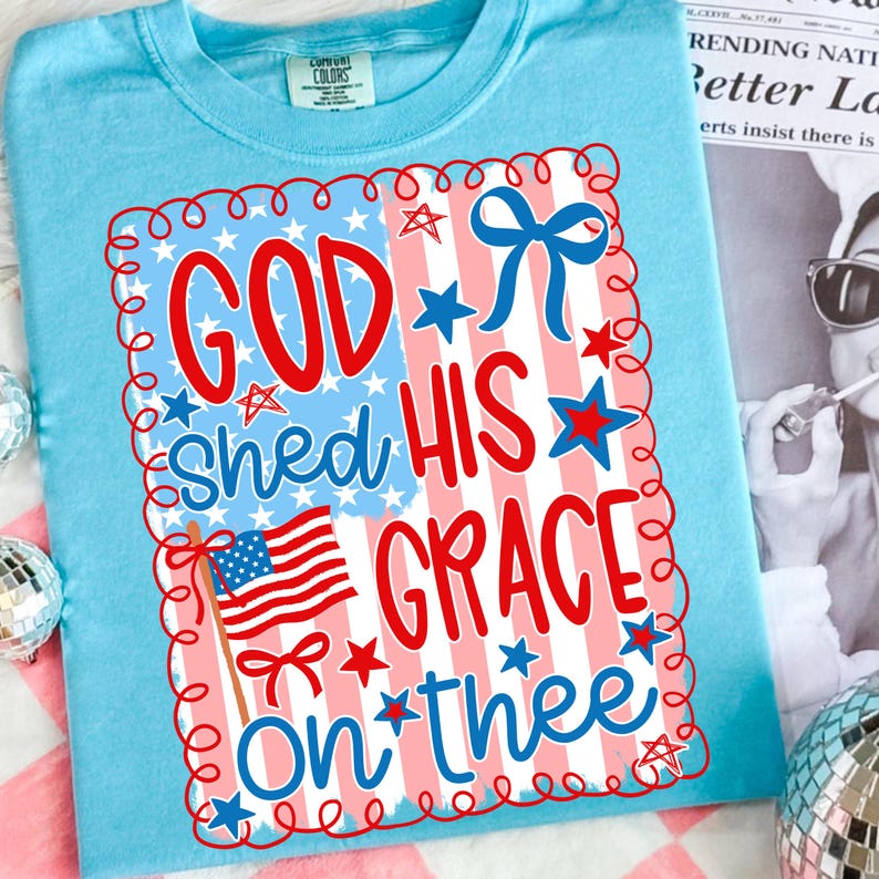 May include: A light blue t-shirt with a patriotic design. The graphic features the text "GOD SHED HIS GRACE ON THEE" in red and blue, along with an American flag and stars. The design is framed with a red and white border.